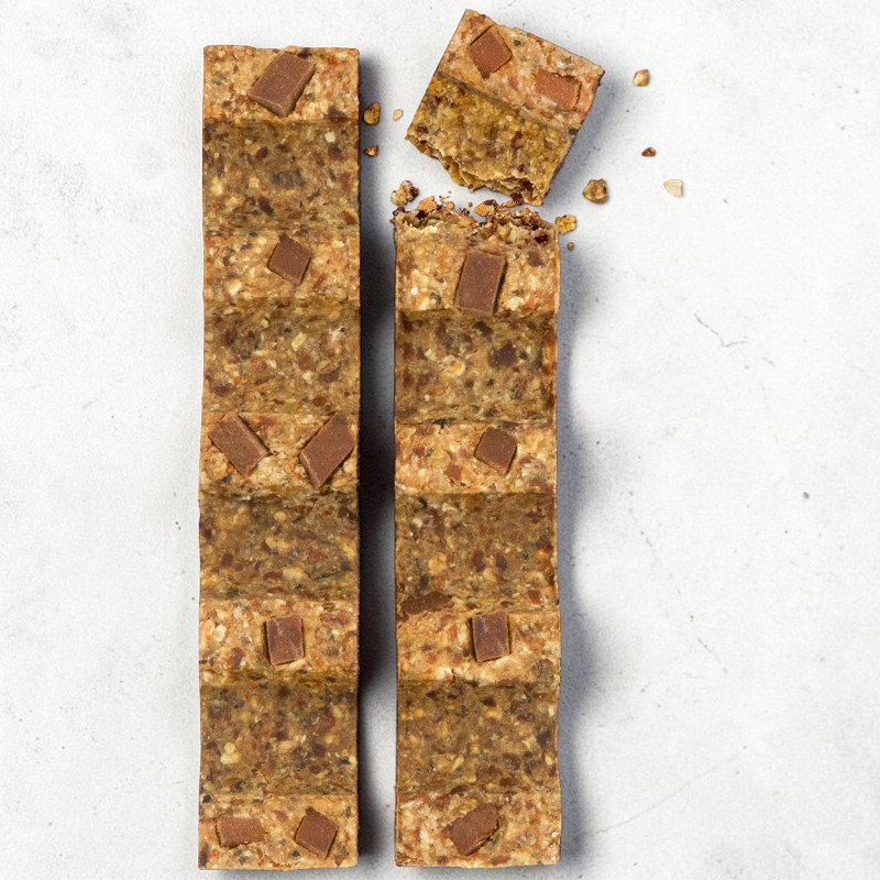 Due barrette allungate su una superficie chiara: la sinistra è intera, la destra è leggermente spezzata in alto con briciole. Entrambe sono di cereali e frutta secca, decorate con pezzetti rettangolari di cioccolato.