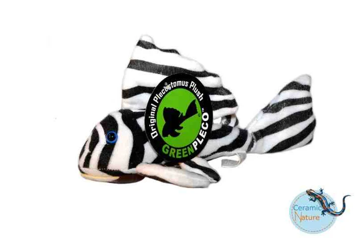 Gestreiftes Plüschtier in Form eines Plecostomus-Fisches auf weißer Oberfläche. Schwarze und weiße Querstreifen, Flossen und Schwanz. Grünes rundes Etikett: "Original Plecostomus Plush", "GREEN PLECO" mit Fisch-Silhouette. Blaue Knopfaugen.
