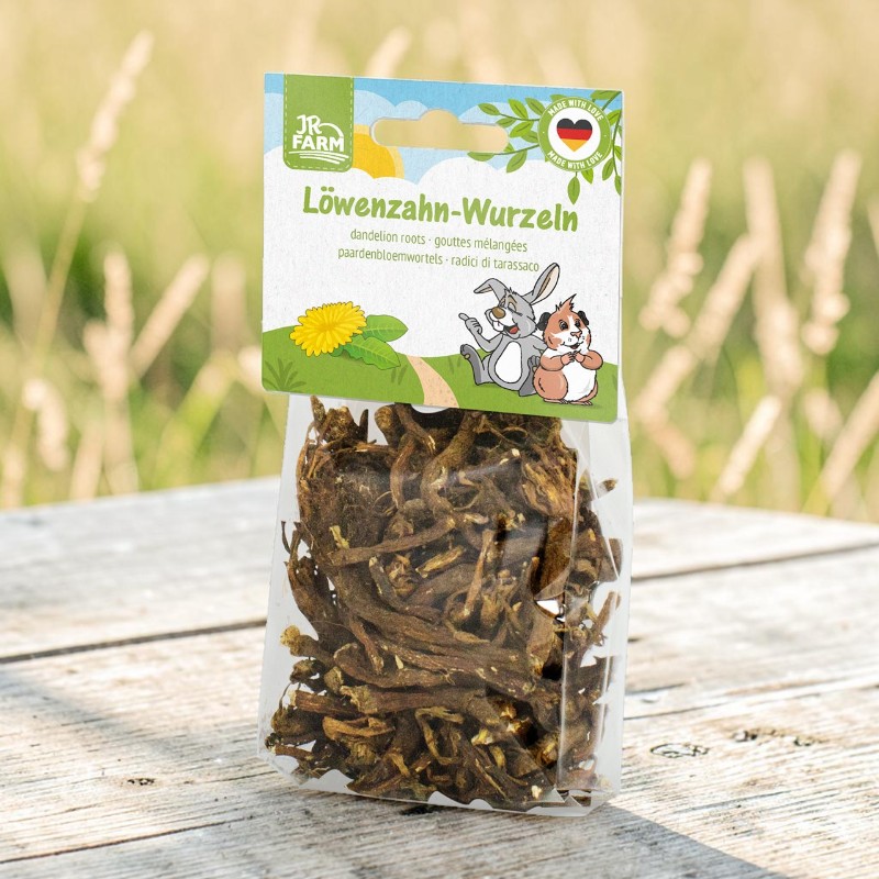 A transparent plastic bag containing dried dandelion roots is standing on a rustic piece of wood. The bag has a colourful label featuring ‘Dandelion Roots’, a dandelion flower, a rabbit and a guinea pig. In the background is a green meadow.