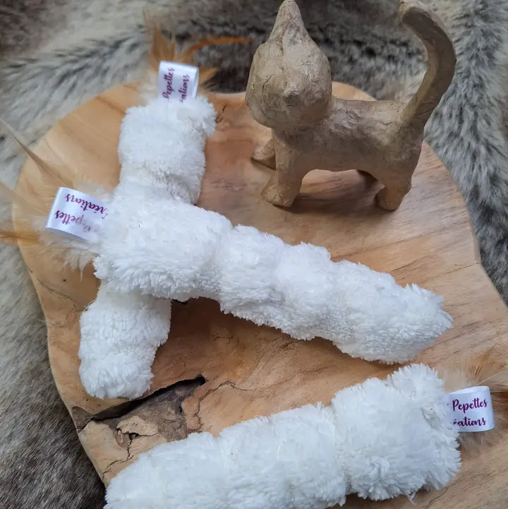Zwei weiße, stoffbezogene Katzenspielzeuge mit Federn und eine Katzenfigur aus Papiermaché auf Holzbrett.