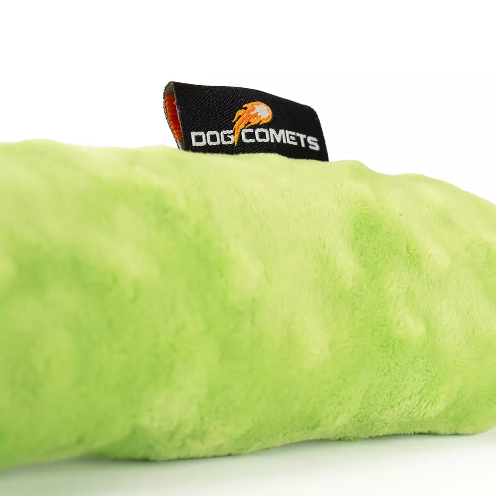 Dettaglio di un giocattolo di peluche per cani di un vivace verde lime dalla consistenza morbida e soffice. Fissata sulla parte superiore: etichetta nera con la scritta bianca "Dog Comets" e il logo a forma di cometa arancione, su sfondo chiaro.