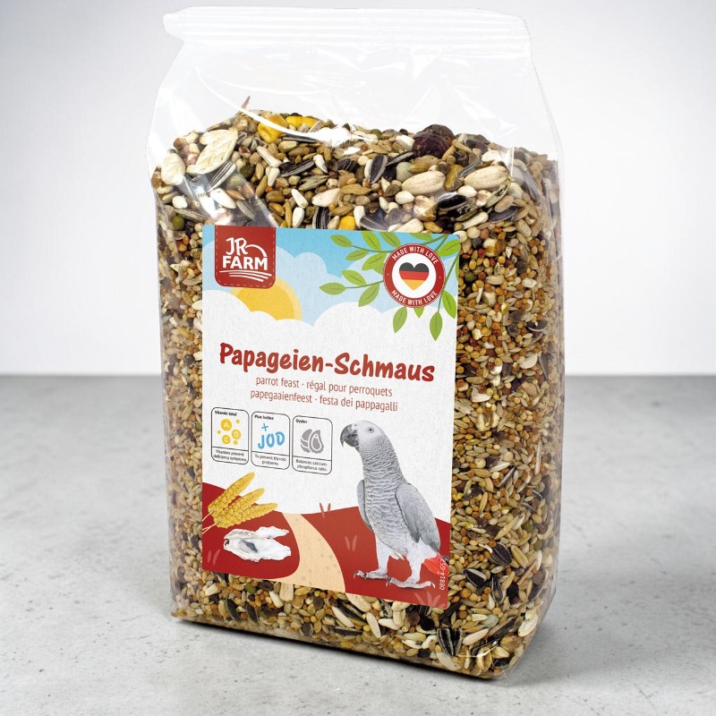 Sachet transparent contenant de la nourriture pour perroquets : mélange de graines de tournesol, de graines de courge, de céréales et de fruits secs. Étiquette colorée : « JR Farm Festin pour perroquets », perroquet gris, épis de blé, vitamines et iode.
