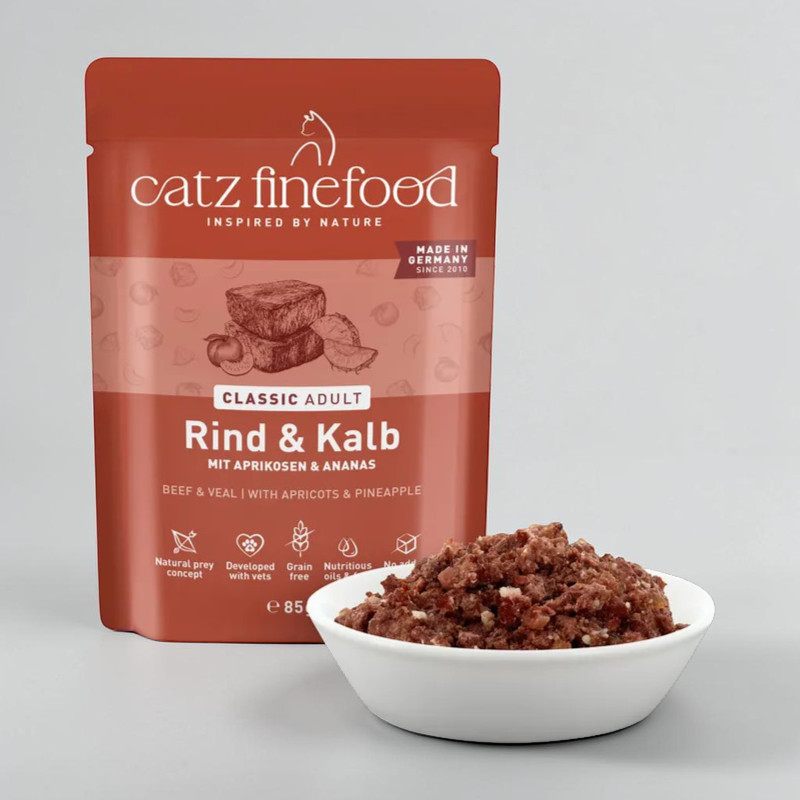 Eine braune catz finefood Futterpackung für Katzen, beschriftet mit "Rind & Kalb mit Aprikosen & Ananas", steht neben einer weißen Schale voller feuchtem, dunkelbraunem Katzenfutter. Die Packung zeigt auch "Classic Adult" und "85g".