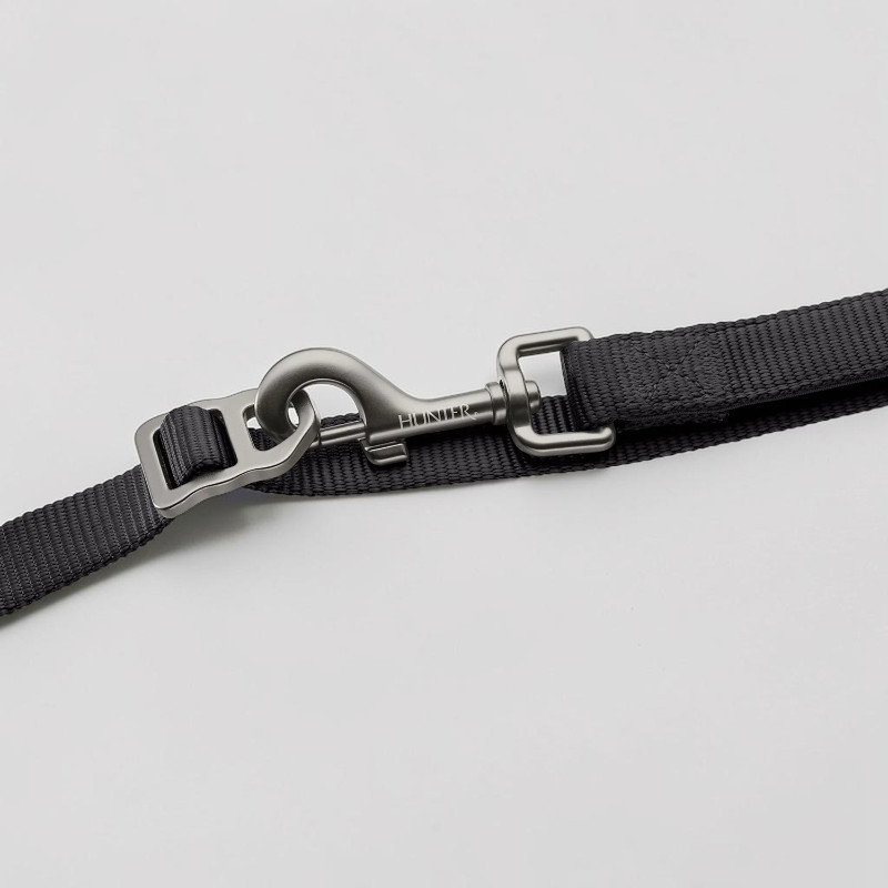 Laisse pour chien noire en sangle tissée robuste, dotée d'un mousqueton de sécurité en aluminium gris mat. Le logo « Hunter » est gravé. Un réglage métallique rectangulaire sur la sangle permet d'ajuster la longueur.