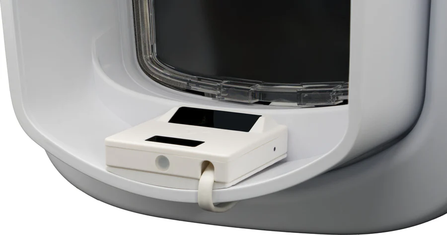Nahaufnahme eines modernen, weißen Haustier-Futterautomaten oder einer Katzentoilette. Sichtbar ist ein transparentes, oben öffnendes Klapptor. Darunter ein kompaktes weißes Sensormodul mit zwei schwarzen Anzeigen, per Kabelband verbunden.