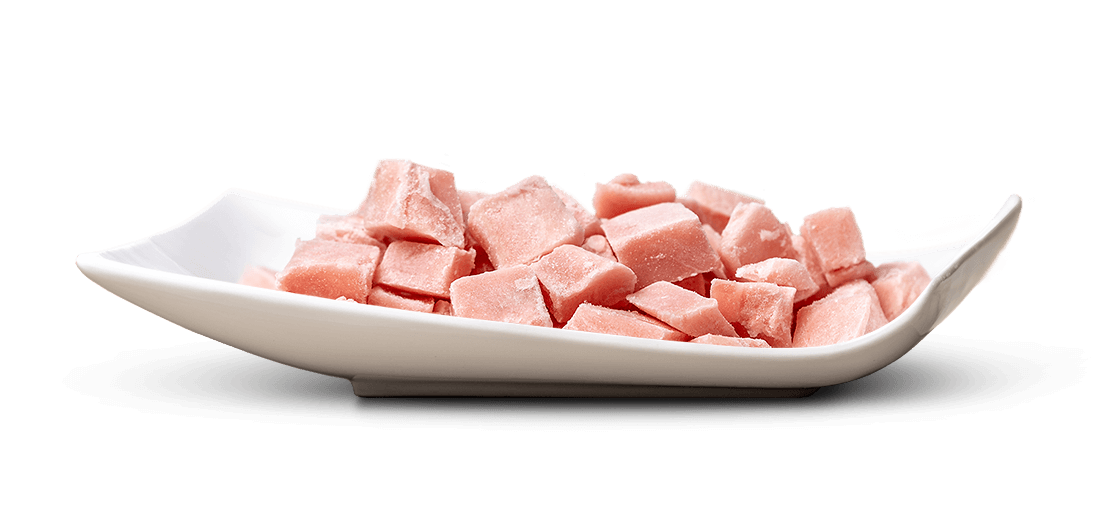 Eine weiße, schalenförmige Schüssel ist mit vielen kleinen, unregelmäßig geformten, rosafarbenen Würfeln gefüllt. Ihre Oberfläche wirkt leicht gefroren oder zuckrig, uneben. Sie liegen eng beieinander.
