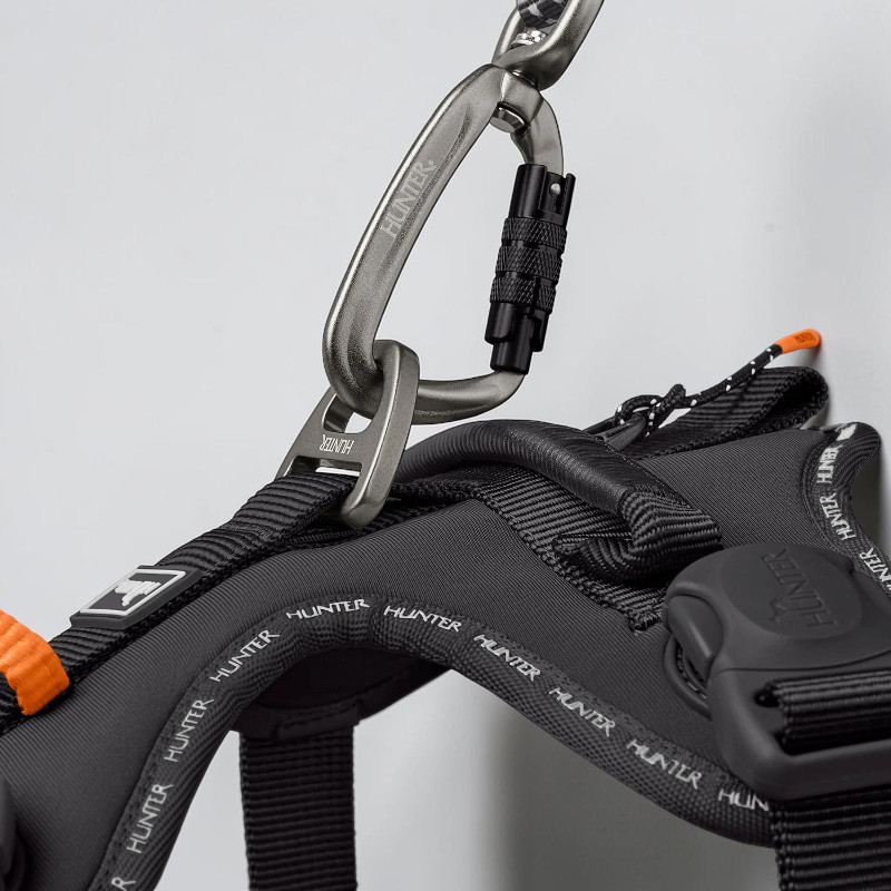 Close-up of the black ErgoMove Pro dog harness: grey snap hook on the sturdy aluminium D-ring. Ergonomically designed with thick padding, black straps, reflective Hunter lettering and a handle.