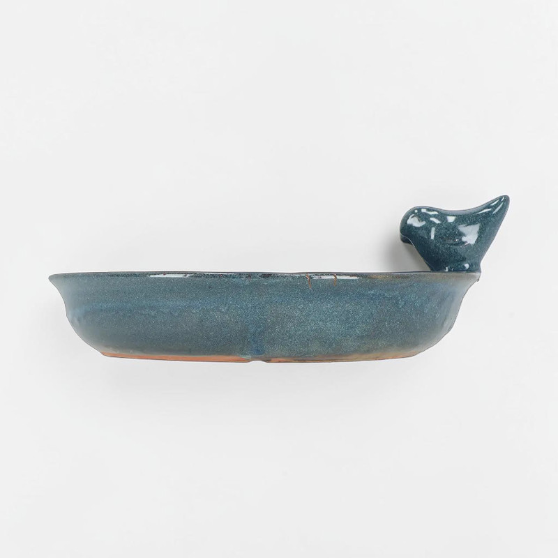 Abreuvoir rond pour oiseaux en céramique brillante couleur pétrole. Bol plat avec large rebord où est perchée une petite figurine d’oiseau stylisée assortie, tête tournée à droite. Dessous non émaillé brun-rougeâtre.