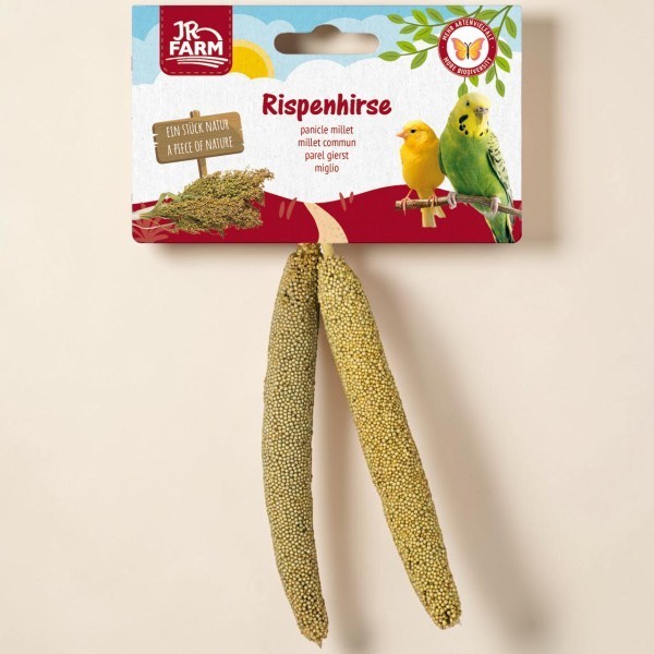 Two long, golden-green panicle millet stalks hang from a colourful ‘ JR Farm ’ packet. It features a blue sky, green leaves and a yellow canary, as well as a green budgie perched on a branch, for natural bird feed.