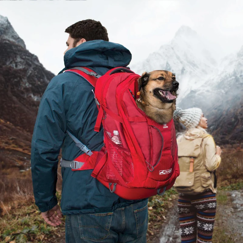 Man with a red dog rucksack against a mountain backdrop. A dog looks out of the rucksack. A woman with a rucksack walks behind.
