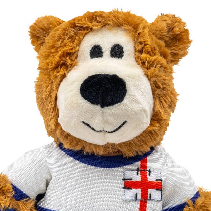 Orsacchiotto marrone di peluche con muso color crema, occhi e naso neri. Indossa una maglietta bianca con colletto blu e una croce rossa ricamata su sfondo bianco, simile alla bandiera inglese. Sorride dolcemente.