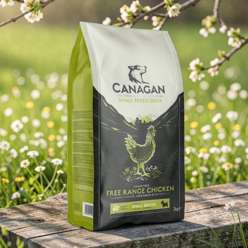Un sachet de croquettes Canagan pour petites races au poulet élevé en plein air est posé sur du bois rustique. L'emballage blanc, noir et vert montre un poulet et des montagnes. Derrière, une prairie fleurie floue et une branche de fleurs blanches.