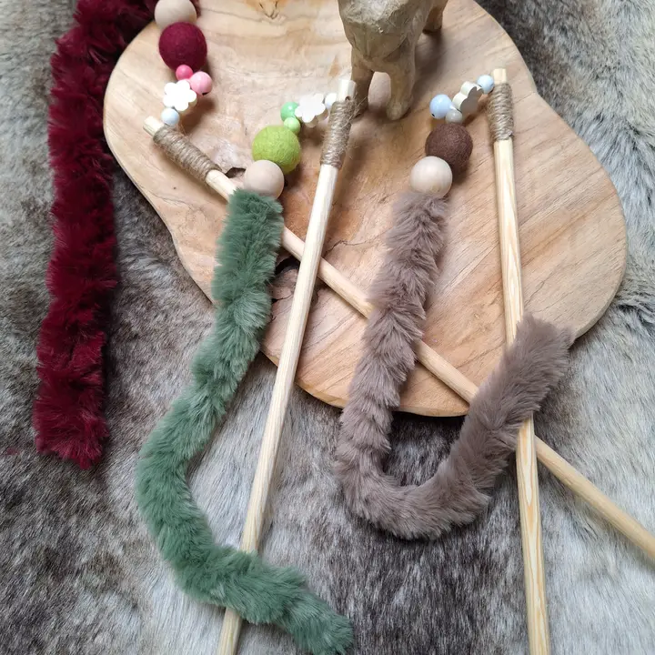 Handgefertigtes Katzenspielzeug: Stöcke mit Plüschschwänzen in Grün, Braun, Rot auf Holzbrett, daneben eine Holzfigur auf Fell.