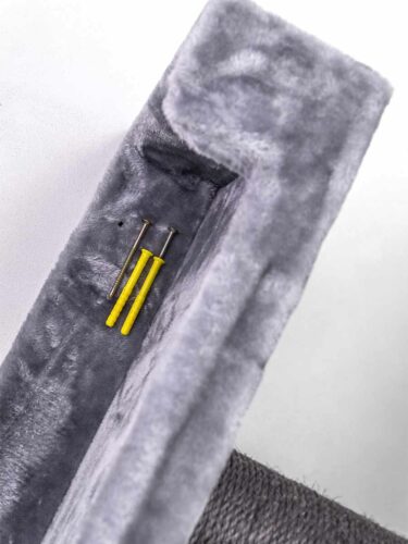 Su una superficie grigia e soffice giacciono due viti argentate e due tasselli gialli per il montaggio. La superficie fa parte di un oggetto a forma di L, forse un elemento di un tiragraffi per gatti. In basso è visibile del materiale scuro intrecciato.
