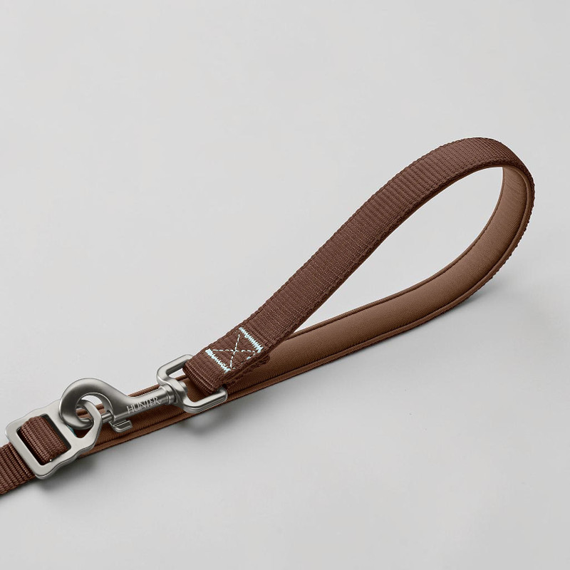 Braune `Action ErgoMove Pro` Hundeleine mit gepolsterter Handschlaufe in hellerem Braun. Silberner Alu-Karabiner mit 'HUNTER'-Gravur. Nahe dem Karabiner X-förmige, hellblaue reflektierende Nähte. Auf hellgrauem Untergrund.