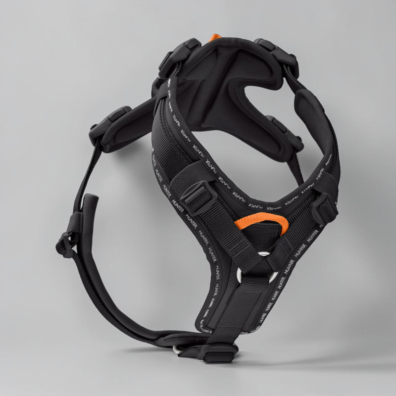 Black ergonomic dog harness on a light grey background. Wide padded straps, Y-shaped chest panel and reflective Hunter lettering. Orange accents on the D-ring and handle, adjustable buckles.