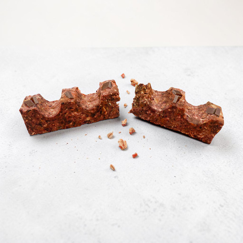 Una barretta di colore rosso-marrone, con superficie ondulata e piccoli pezzetti di cioccolato, è spezzata a metà su uno sfondo grigio chiaro. Le due metà sono leggermente distanziate l'una dall'altra, con briciole e pezzetti nel punto di rottura.