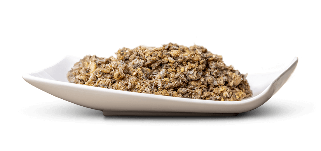 A white rectangular dish with curved edges, filled with a coarsely chopped, grainy mixture of irregular pieces in shades of dark brown, grey and beige, resembling tapenade or a spicy mushroom paste, set against a white background.