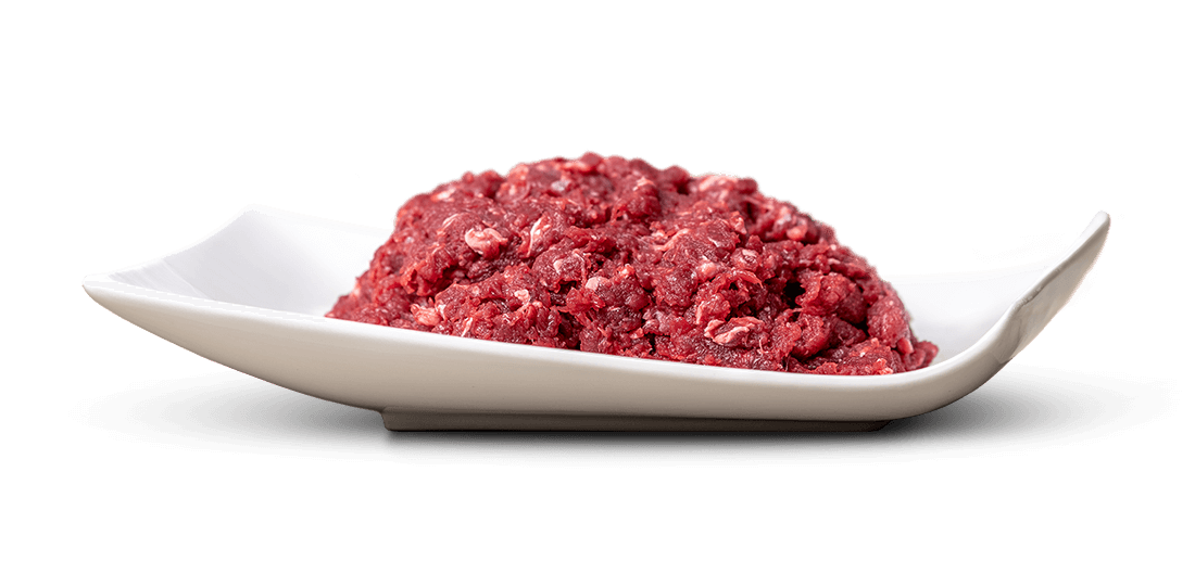 Un tas de viande hachée rouge, parsemé de petits morceaux de graisse blanche, est disposé au centre d'un plat rectangulaire blanc et plat, aux bords légèrement surélevés. Le plat repose sur un fond clair qui ne projette qu'une ombre très légère.