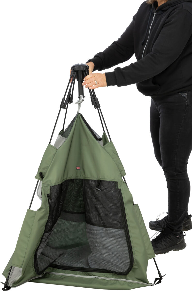 Una persona con felpa con cappuccio scura e jeans monta una tenda per animali domestici verde oliva, conica, su sfondo bianco. La tenda ha finestre in rete nera e la persona tiene il meccanismo superiore. Logo Trixie visibile.