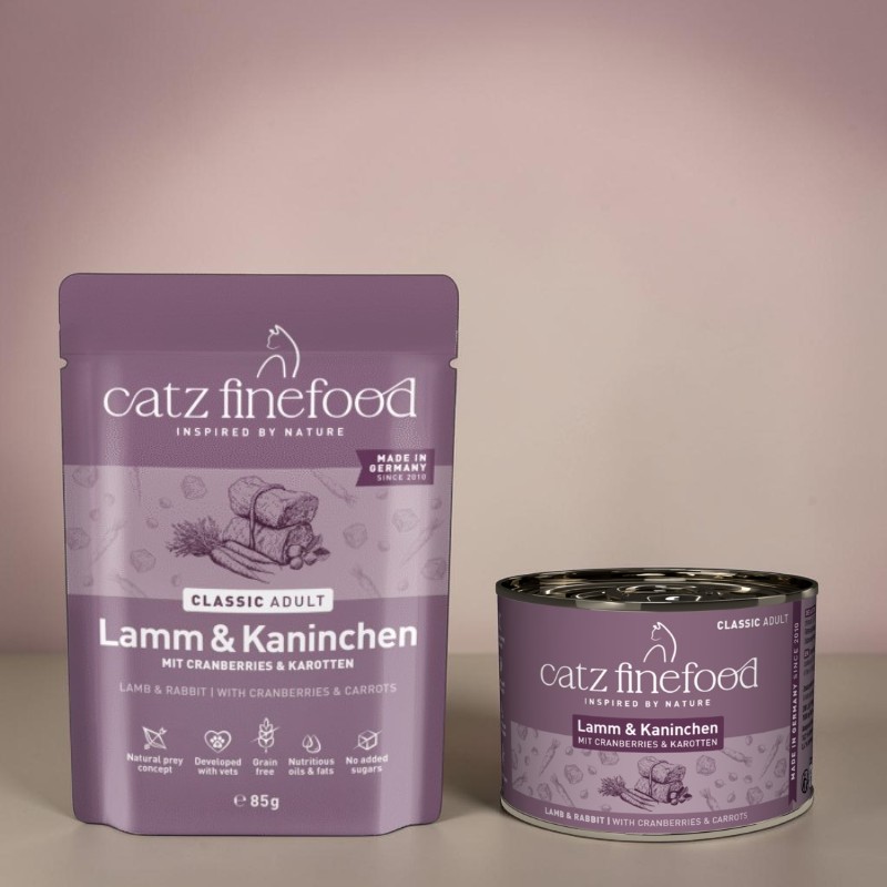 Packaging viola da esposizione per il cibo per gatti «Catz Finefood Agnello e coniglio con mirtilli rossi e carote» su sfondo chiaro. Accanto, barattolo viola abbinato con etichetta e informazioni. Per gatti adulti, senza cereali.
