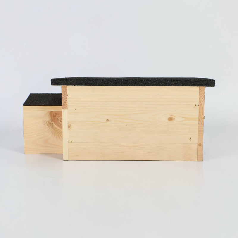 Maisonnette pour hérissons en bois d'épicéa clair avec un toit en bitume foncé, photographiée de profil. Structure allongée en bois comprenant une pièce principale et une petite entrée décalée sur la gauche. Veines et nœuds visibles. Abri protégé.