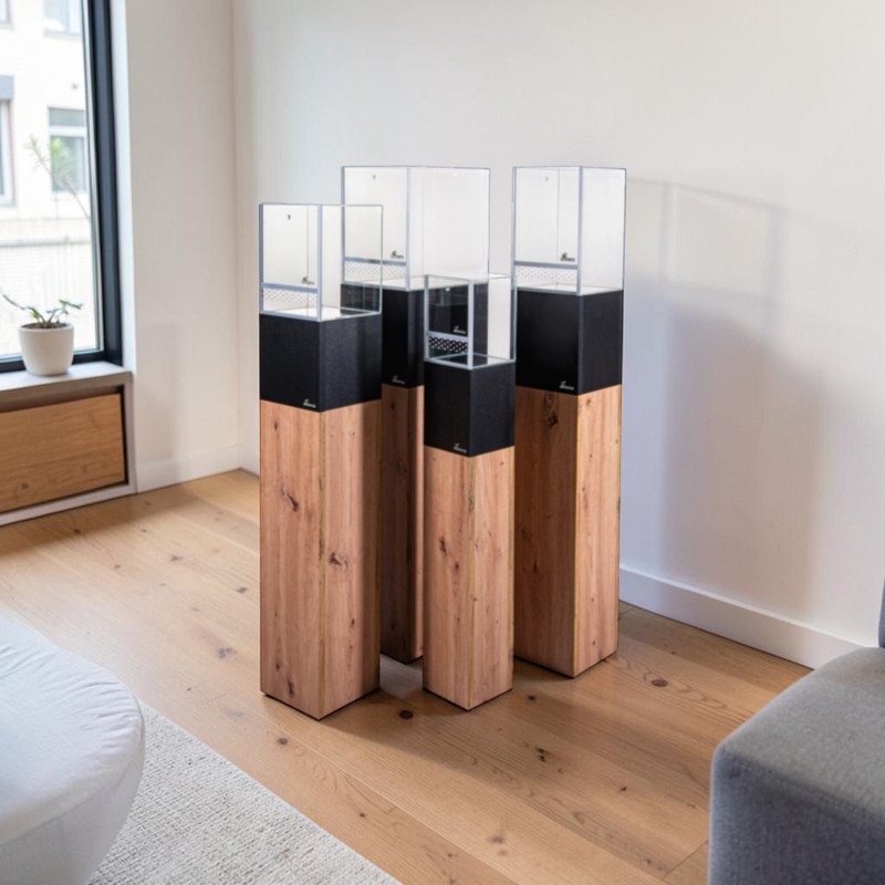 Four modern, rectangular display columns of varying heights, with light brown wooden bases, black central sections and transparent acrylic tops. Set on a wooden floor in a bright room by the window, with a building in the background.