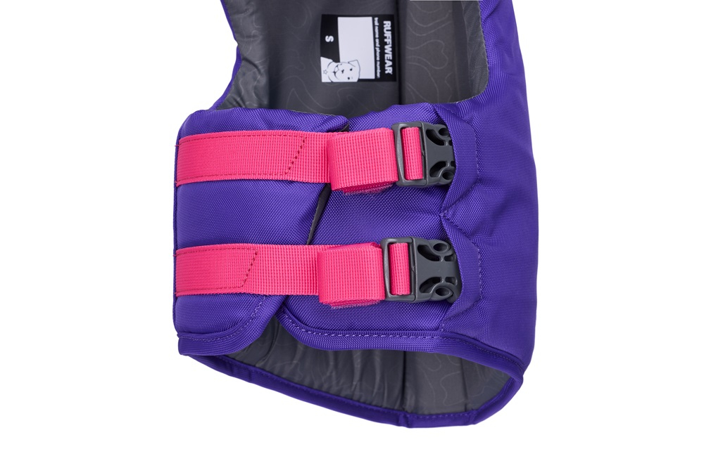 Gilet de sauvetage violet pour chien, avec deux sangles rose vif et boucles noires, horizontales sur la poitrine. Étiquette noire « Ruffwear » en haut, taille « S », doublure grise à motifs. Légèrement de profil, vu de face.