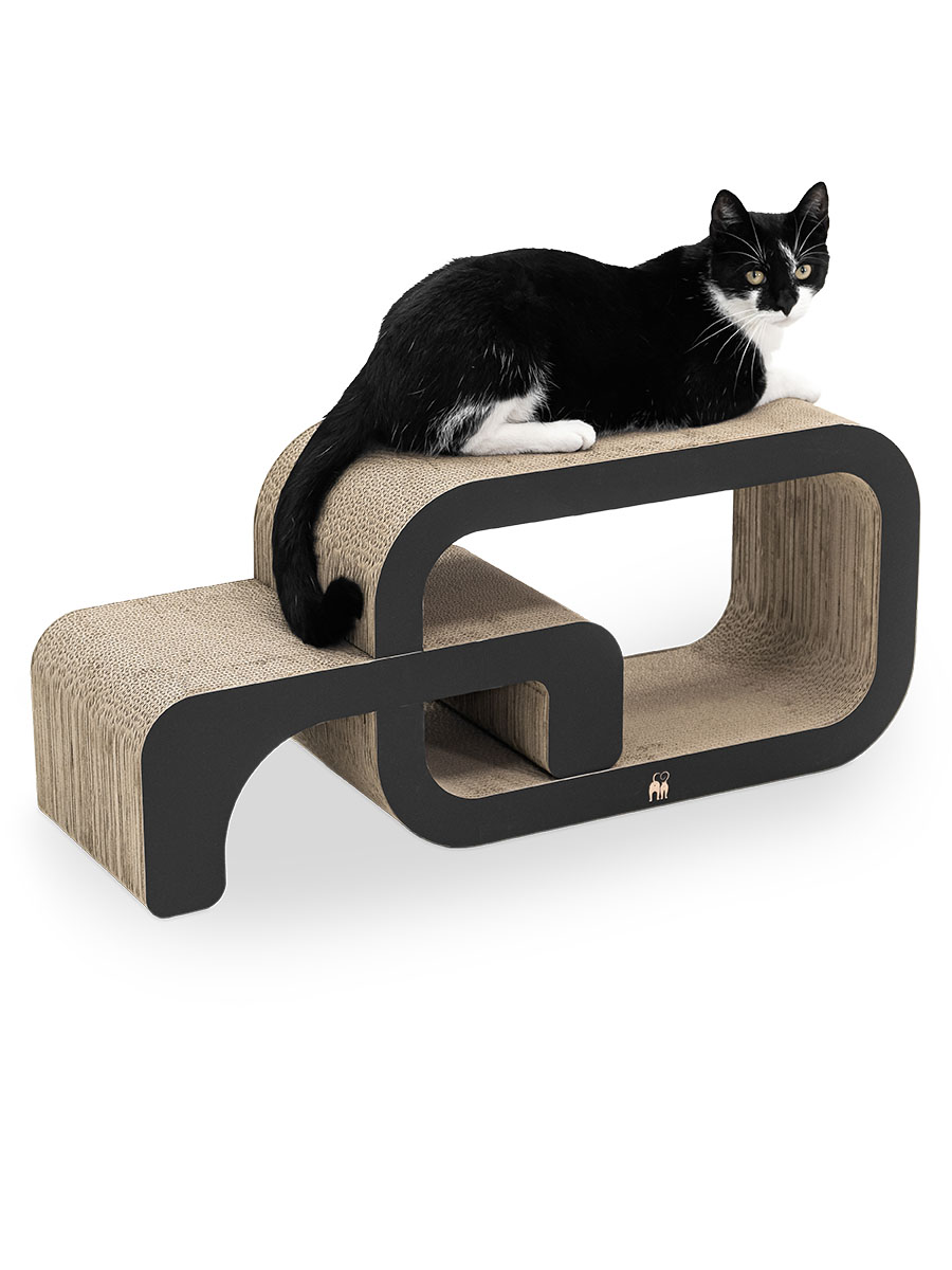 Un gatto bianco e nero giace rilassato su un moderno tiragraffi a due livelli realizzato in cartone ondulato. Il design sinuoso con bordi neri sembra una forma astratta a S. Il gatto guarda in avanti.