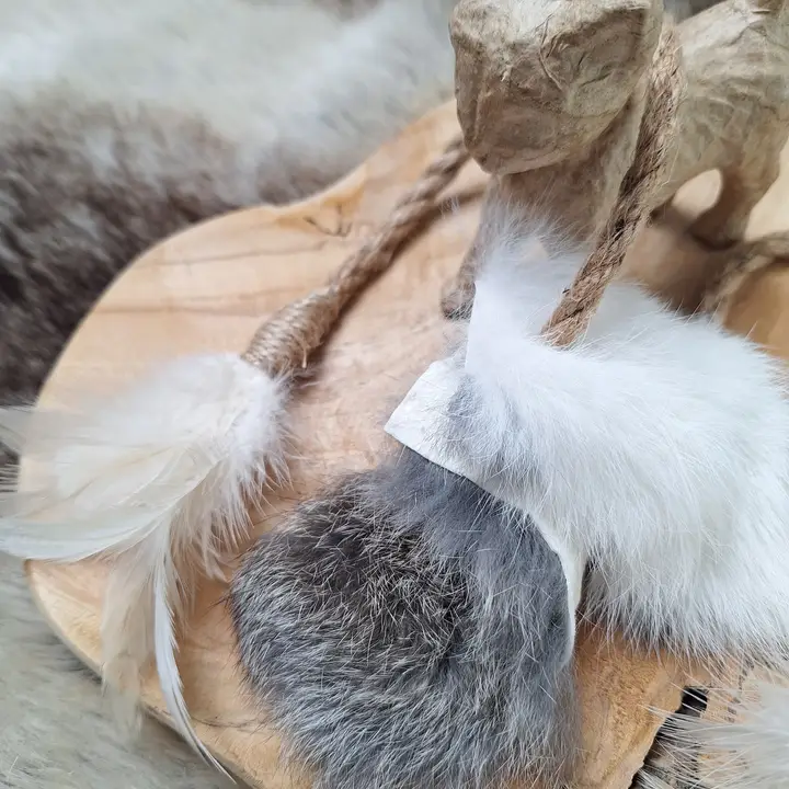 Dekorative Anordnung aus Holz, Papierfigur, Seil, Federn und Fell auf einem Fellhintergrund.