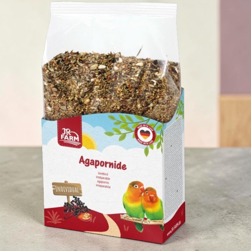 Transparent bag of bird feed in a colourful cardboard sleeve. Label: ‘JR Farm’ and ‘Lovebirds’ above two green-and-orange lovebirds on a branch. Also a ‘Made with Love’ seal (German flag) and an ‘INDIVIDUAL’ wooden sign with elderberries.