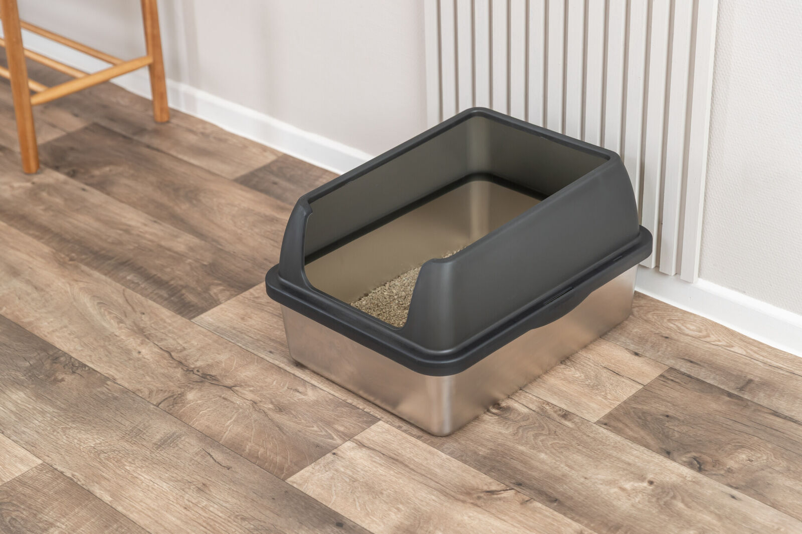 A cat litter tray with a silver-coloured base and a dark grey plastic top with an arched opening. Light brown cat litter can be seen inside. It stands on a light brown wooden floor next to a white wall with decorative panelling.