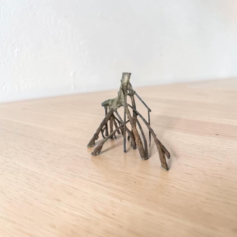 Detailreiche Struktur wie freiliegende Wurzeln oder ein Miniatur-Baumstumpf mit vielen dünnen, spinnenartigen Stützen auf heller Holzoberfläche vor schlichter weißer Wand. Oberfläche uneben, dunkel-erdige Töne.