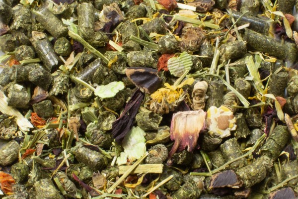 Nahaufnahme einer Mischung aus groben, trockenen Tierfutterpellets in Grüntönen, mit hellgrünen geriffelten Blättern, rötlich-braunen Blütenblättern, dünnen hellgrünen Grashalmen und kleinen orangefarbenen Stückchen.