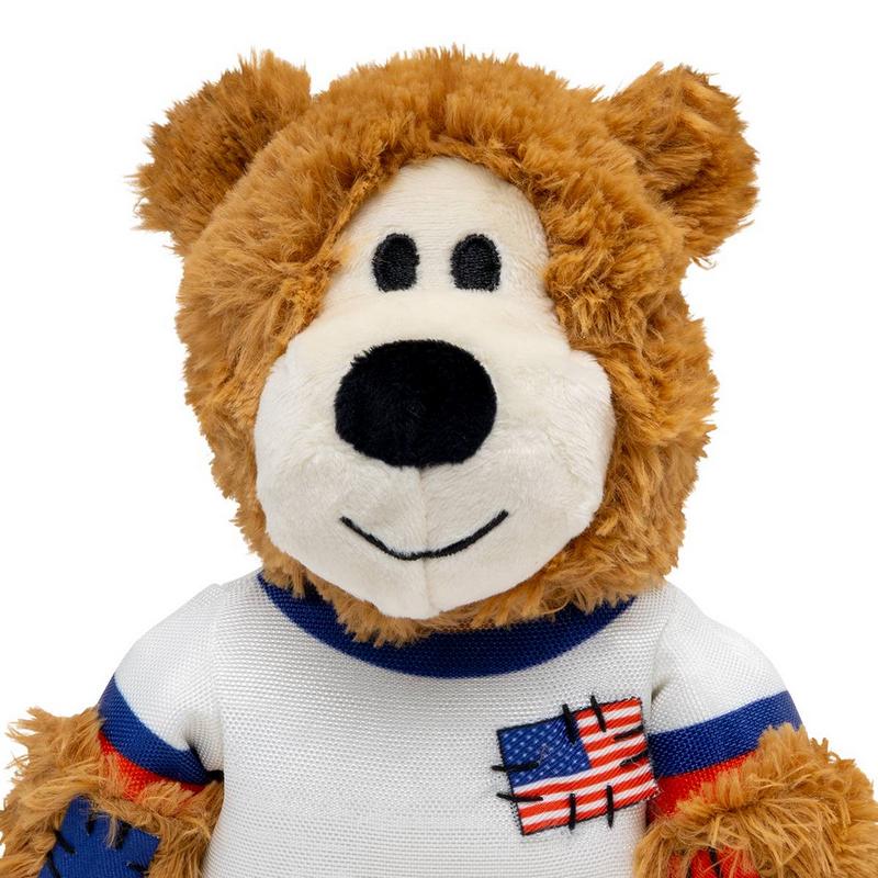 Morbido orsacchiotto marrone dal viso simpatico. Indossa una maglietta bianca con strisce rosse e blu sulle maniche e un distintivo ricamato con la bandiera degli Stati Uniti. Bocca bianca, naso nero in peluche, sorriso ricamato.