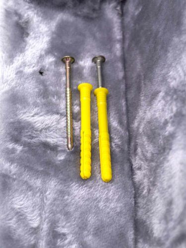 A sinistra un chiodo in metallo argentato con gambo zigrinato. A destra due tasselli in plastica giallo brillante, ognuno con un chiodo in metallo argentato parzialmente inserito. Su sfondo morbido grigio tipo pile.