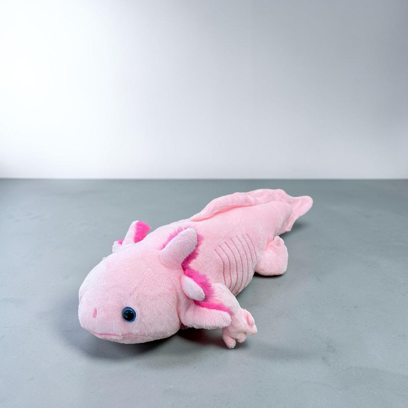 Ein weiches, pinkes Axolotl-Plüschtier liegt auf einer schlichten grauen Oberfläche. Es hat leuchtend blaue Augen und fächerartige, pinke Kiemenbüschel an den Kopfseiten. Im Hintergrund eine neutrale weiße Wand.