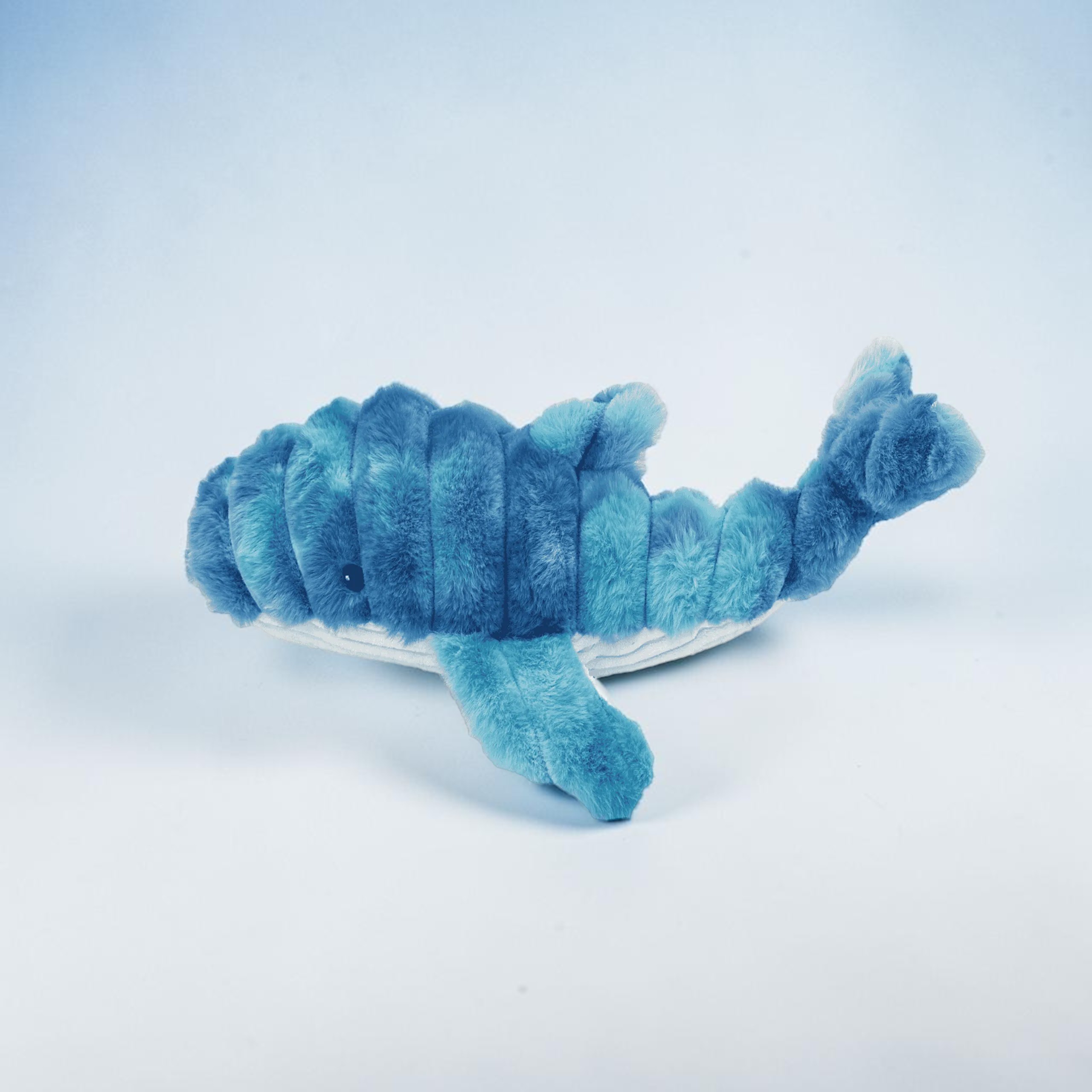 Ein flauschiges, blaues Stofftier in Form eines Wals liegt auf einer hellblauen Oberfläche. Der Wal hat einen strukturierten, dunkleren Rücken und hellblaue Flossen und Schwanz. Sein Bauch ist weiß und es ist ein kleines, schwarzes Auge zu erkennen.