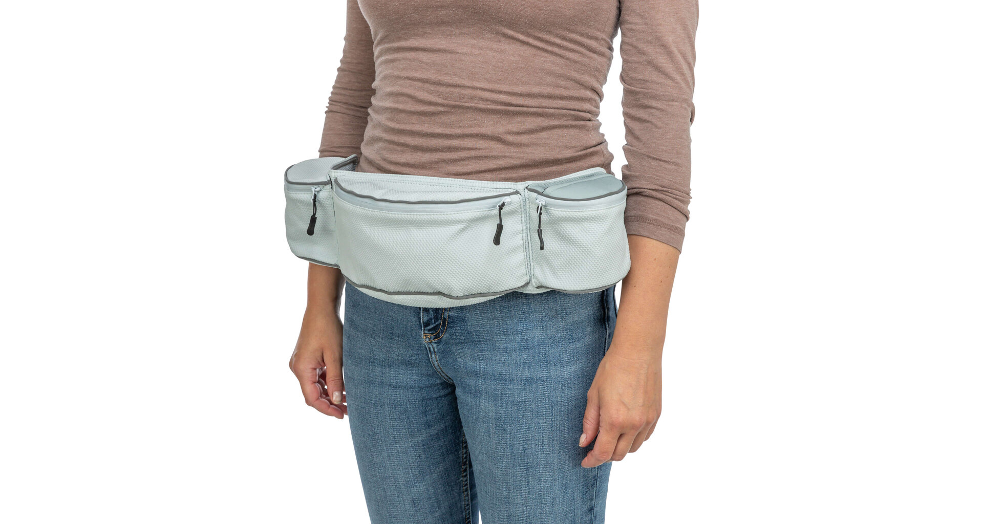 Eine Person, vom Oberkörper bis zu den Oberschenkeln, steht vor weißem Hintergrund. Sie trägt ein hellbraunes Langarmshirt und blaue Jeans. Um die Taille eine hellblaue Bauchtasche mit Fächern, Reißverschlüssen und grauen Akzenten.