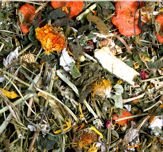 A detailed close-up of a natural mixture of dried herbs, grasses, pieces of root and plant parts. Green and brown leaves, yellowish flowers, pieces of carrot and light-coloured roots can be seen.