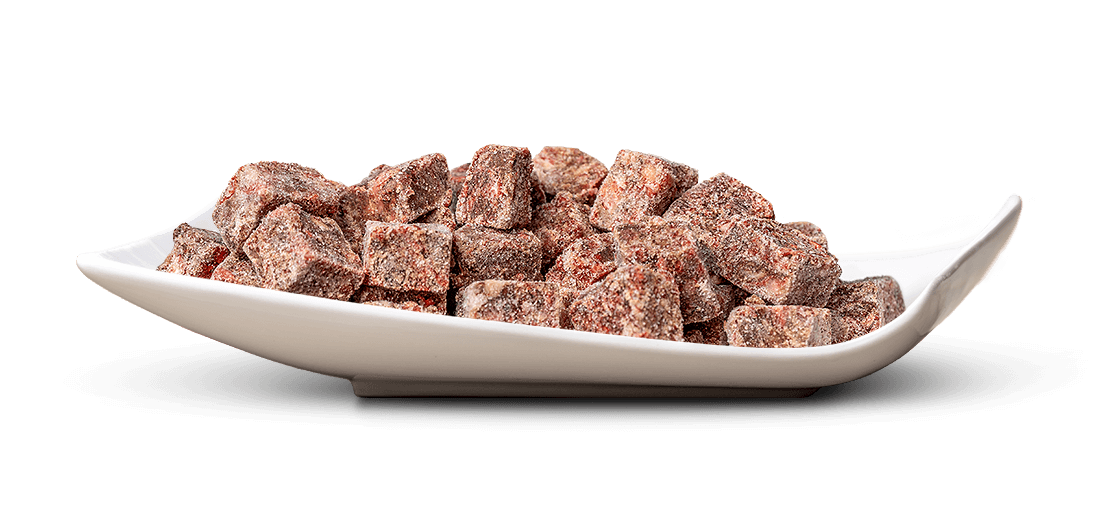 Kleine, unregelmäßige, rötlich-braune Würfel mit feiner heller Gewürz- oder Zuckerschicht liegen auf einer elegant geschwungenen weißen Schale. Raue, trockene Textur, appetitlich angeordnet. Rein weißer Hintergrund.