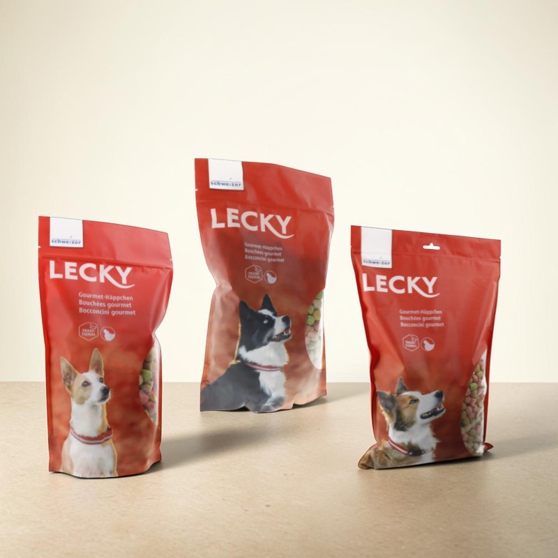 Trois sachets rouges de friandises pour chiens « Lecky » sont posés sur une surface claire. Chaque sachet montre une tête de chien et, sur le côté, des biscuits croustillants : gauche marron/blanc ; centre noir/blanc ; droite marron/blanc.