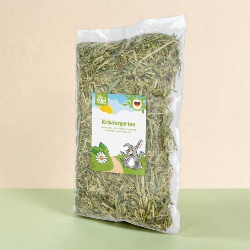A transparent plastic bag, filled with dried, coarsely chopped green hay or a herb mixture, standing upright. The ‘JR Farm Herb Garden’ label is visible, with a smiling rabbit, a field of daisies and the German flag below it.