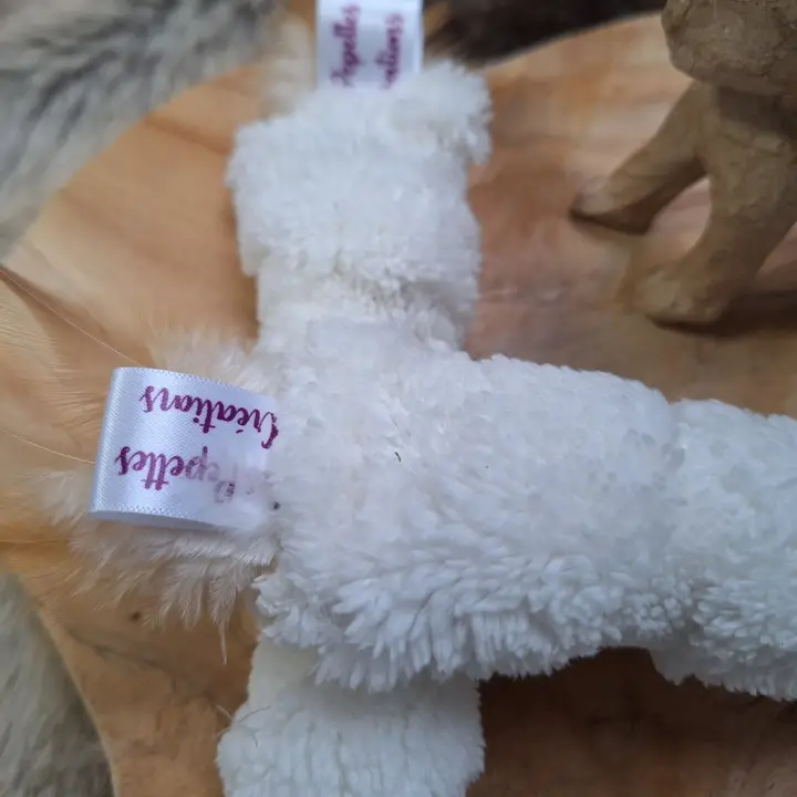 Ein weißes Stofftier mit Etiketten auf einem hölzernen Untergrund, teilweise von Fell umgeben.