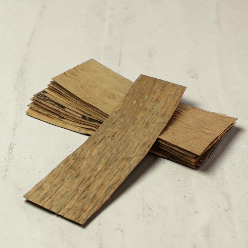 Mehrere längliche, hellbraune Holzstücke mit dunkler Maserung liegen auf einem hellgrauen Untergrund. Ein einzelnes Stück ist im Vordergrund horizontal ausgerichtet, dahinter ist ein Stapel weiterer gleicher Holzscheiben leicht schräg angeordnet.
