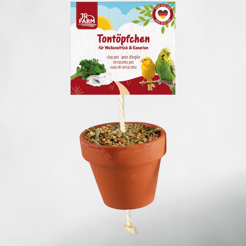 A clay pot filled with bird feed hangs from a cord. Above it is a JR Farm product label reading ‘Clay pots for budgies & canaries’, featuring budgies, canaries, parsley, cuttlebone and the words ‘Made with Love’.