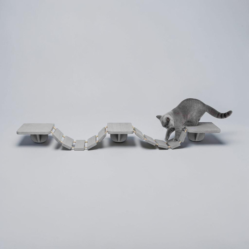 Un chat gris avance prudemment sur une échelle d'escalade modulaire à fixer au mur, de couleur taupe. Des marches en bois et des cordes en sisal forment une structure semblable à un pont suspendu qui relie trois plateformes.