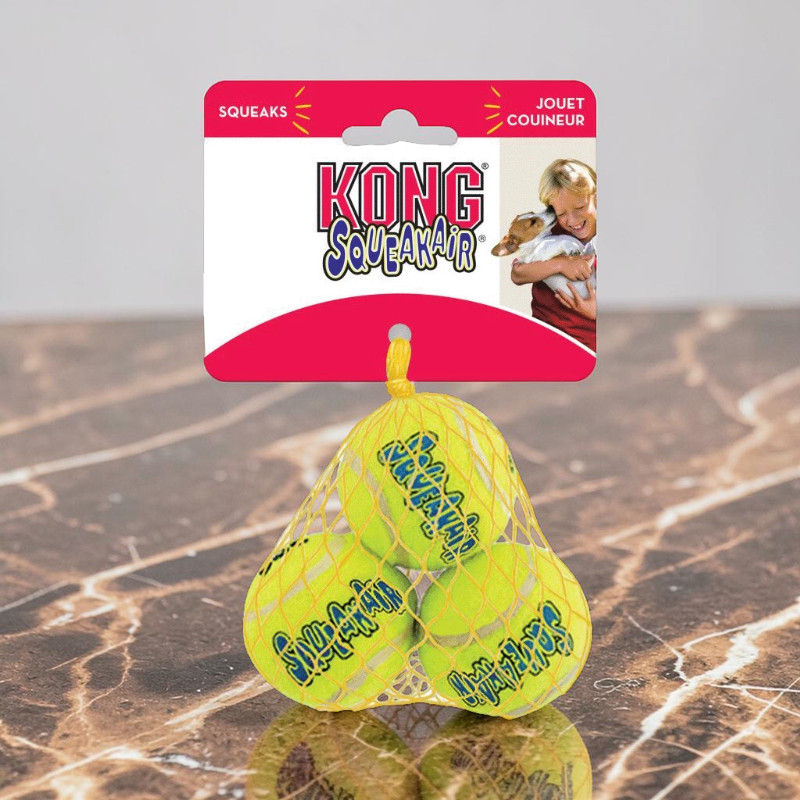 Drei leuchtend gelbe KONG SqueakAir Tennisbälle im gelben Netz an rotem Kartonhänger. Jeder Ball trägt den blauen Schriftzug „SqueakAir“. Der Hänger zeigt KONG Logo, „SQUEAKS“ und ein Kind mit Hund.