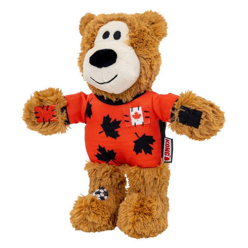 Flauschiger brauner Stoffbär mit schwarzem Gesicht und Ohren. Er trägt ein leuchtend orangefarbenes T-Shirt mit schwarzen Ahornblättern und kleinem Kanada-Flaggenpatch. Vorderpfoten mit roten und schwarzen Patches, eine Hinterpfote mit Fußballmuster.
