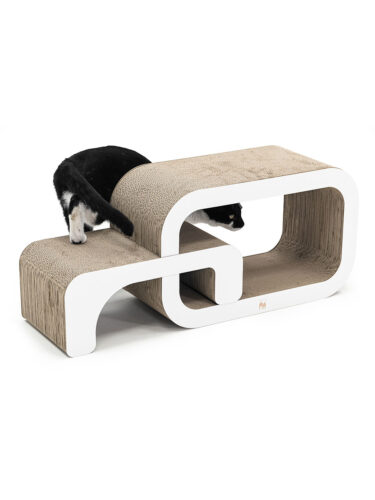 Eine schwarz-weiße Katze spielt mit einem modernen, geschwungenen Kratzmöbel aus Wellpappe mit weißen Rändern. Der hintere Teil der Katze mit dem Schwanz ist links sichtbar, wie sie aufsteigt, während ihr Kopf aus einer Öffnung rechts im Möbel hervorlugt.